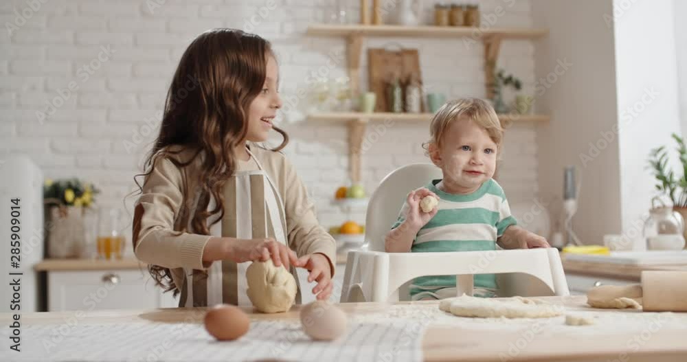Little girl is learning to cook, preparing cake dough while her little brother in baby chair with floured face is playing with piece of pastry - togetherness, childhood dream Little girl is learning to cook, preparing cake dough while her little brother in baby chair with floured face is playing with piece of pastry - togetherness, childhood dream