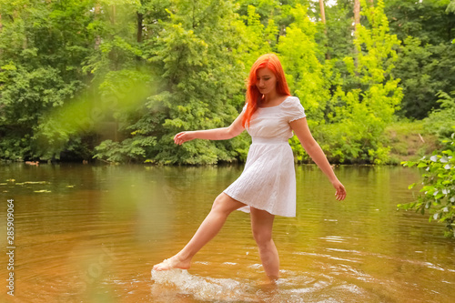 happy redheaded woman in white dress at a river having fun and splashing water in summer. happy person plays with water 