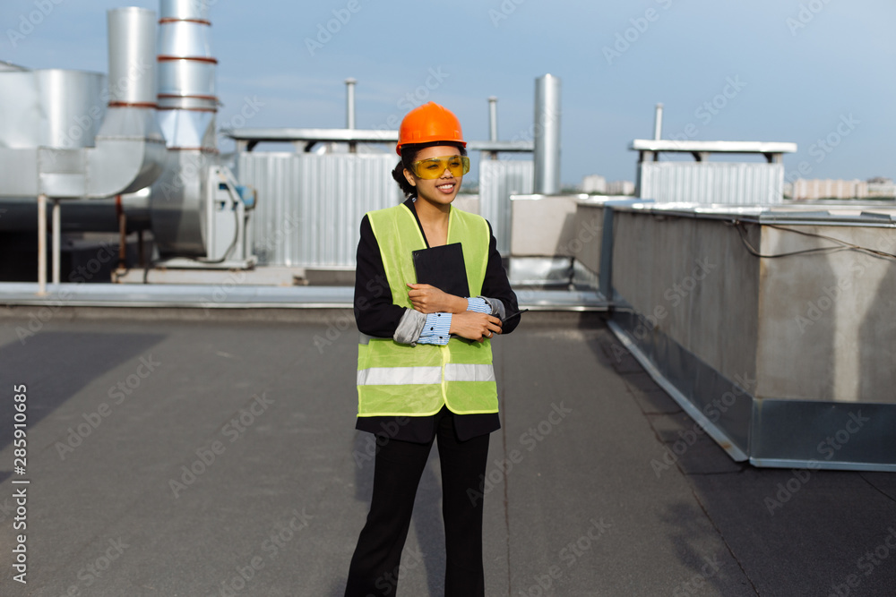 Attractive woman African ethnic young woman at construction site holding a tablet on the rooftop of construction site wearing orange helmet and yellow safety glasses Attractive woman African ethnic young woman at construction site holding a tablet on the rooftop of construction site wearing orange helmet and yellow safety glasses