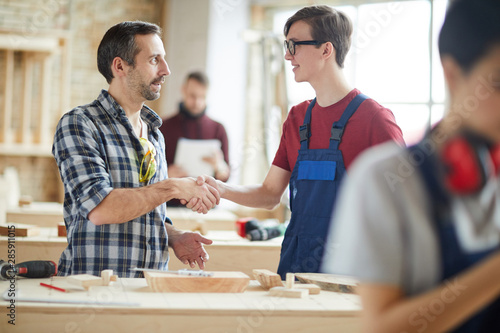 Waist up portrait of mature carpenter shaking hands with young trainee in workshop, copy space