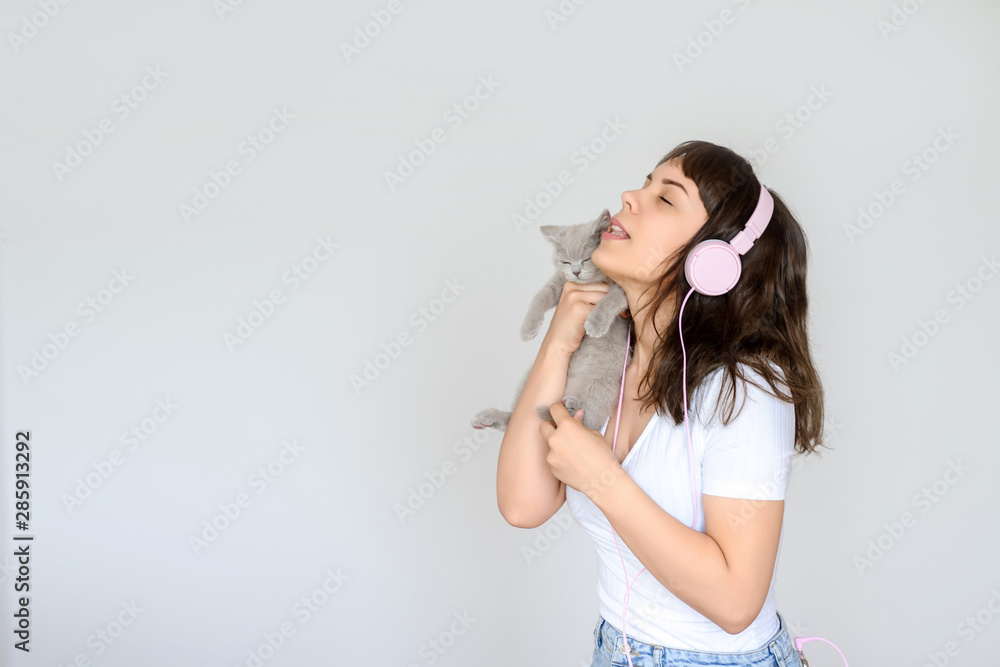 close up photo of a happy brunette with pink headphones listening to the music while holding a gray kitten in her arms close up photo of a happy brunette with pink headphones listening to the music while holding a gray kitten in her arms