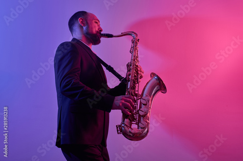 Portrait of professional musician saxophonist man in suit plays jazz music on saxophone  pink background in a photo studio  side view