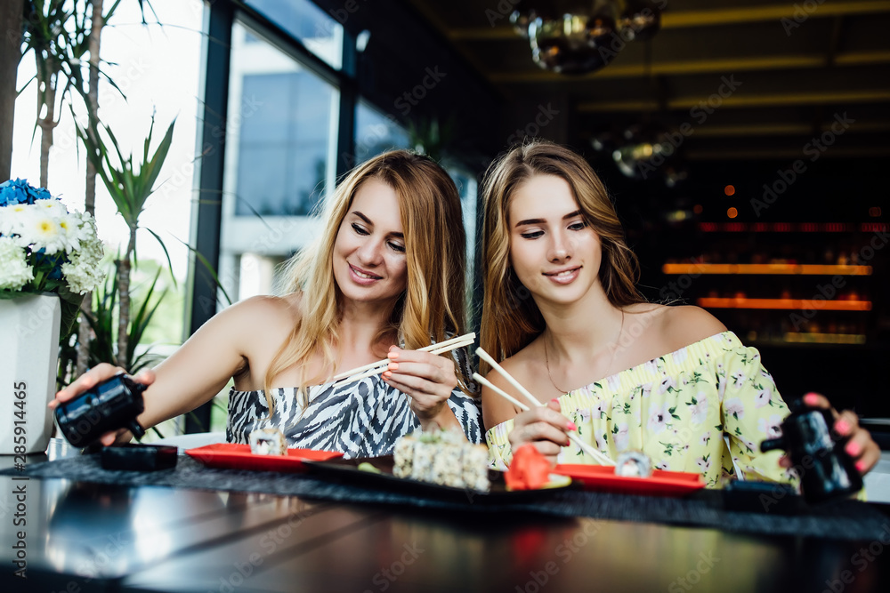 Fototapeta premium Lunch at a Chinese restaurant on the summer terrace. Mom and her young beautiful daughter eat sushi with chinese sticks. Lifestyle.