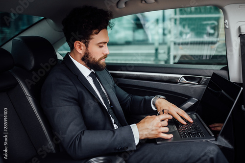 Handsome, bearded, smiling top manager in black suit working on his laptop on the backseat of the car. © Тарас Нагирняк