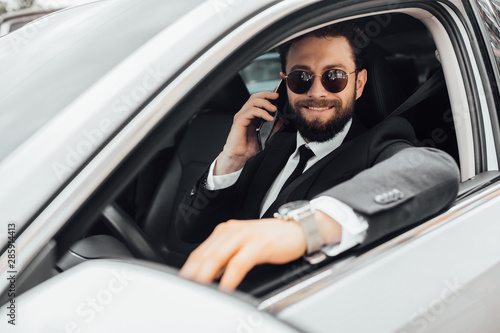 Handsome, bearded, smiling businessman in black suit calling on the phone At the wheel of a white car. © Тарас Нагирняк