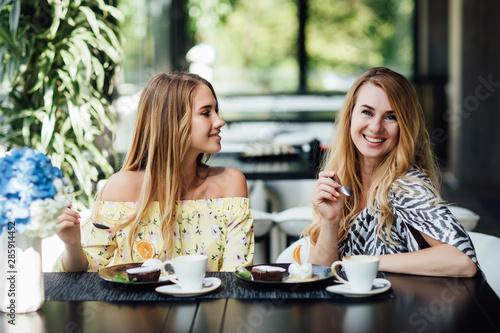 Portrait of mother and blonde daughter talk and smile while drink latte and eating dessert. Spend time happy. Morning breakfast sweets.