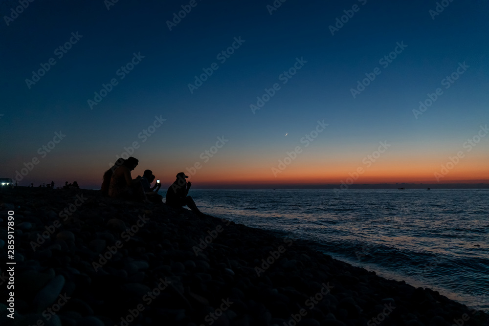 Silhouette of a group of friends who sit on the beach near the sea in the last minutes of sunset, beautiful blue-orange sunset