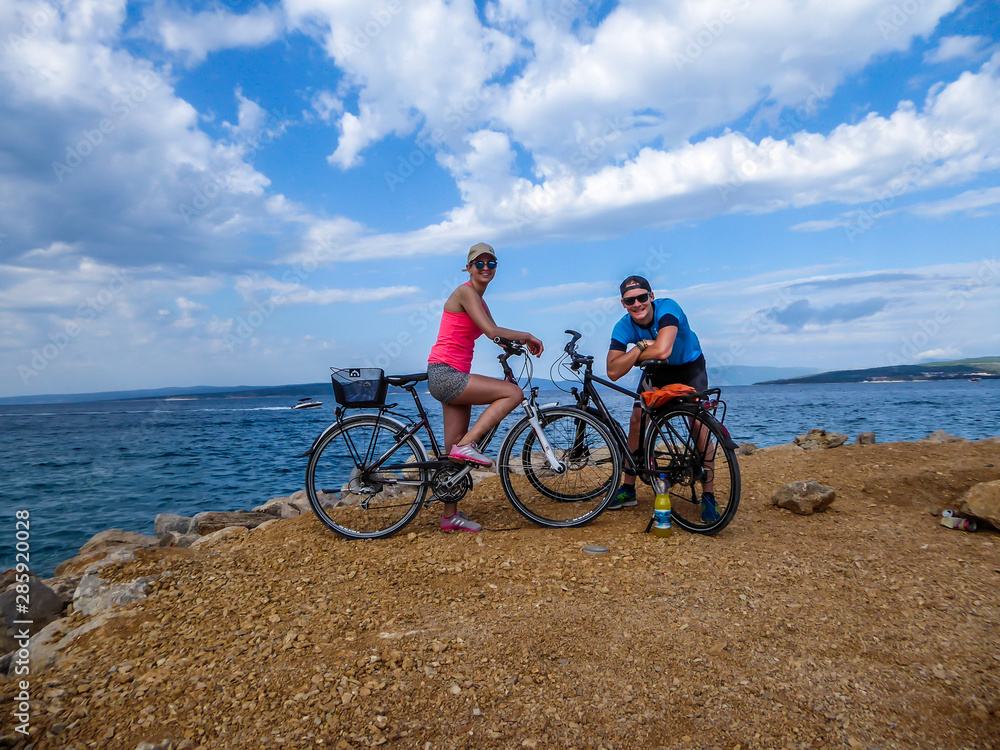 A couple in sporty outfits, standing on their bikes and checking the weather. Behind them there is Mediterranean Sea. Thick clouds above them. Active holidays. Beautiful coastal line of Croatia.