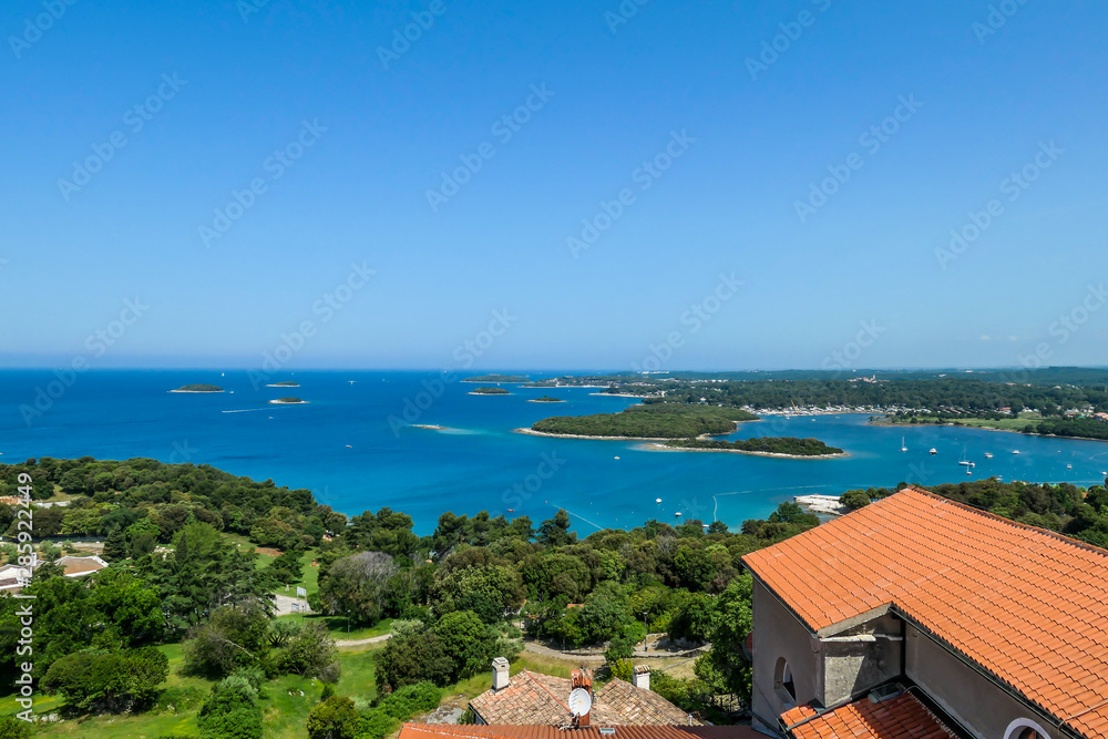 A remote view on the harbour from a hill. Rows of docks waiting for the boats and yachts to anchor there. Lots of houses surrounding the port. In the back there are few islands. Lush green plants