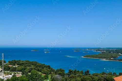 A view on the sea from a little hill. There are plenty small island around the coastal line, each of them overgrown with trees and bushes. There are few boats crossing the sea. Clear and sunny day.