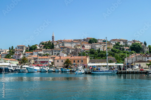 A view on the harbour in Vrsar. There are a lot of boats anchored to the shore. In the back, there is the city center, with a tall church tower. Clear and sunny day. Busy day in the harbour.