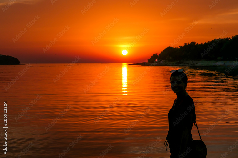 A girl enjoying the sunset by a beach. The sun sets over the horizon. The sun beams reflecting in the calm sea waters. There is an island on the side. The sky turns yellow. Girl enjoys the spectacle