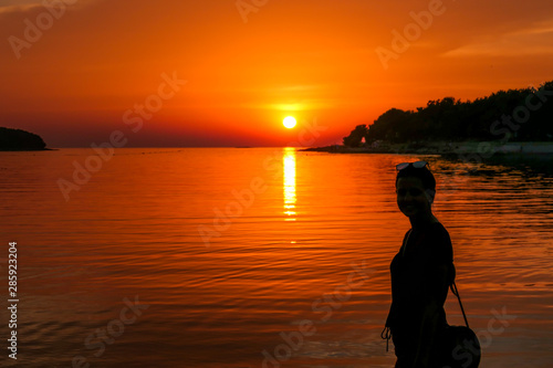A girl enjoying the sunset by a beach. The sun sets over the horizon. The sun beams reflecting in the calm sea waters. There is an island on the side. The sky turns yellow. Girl enjoys the spectacle