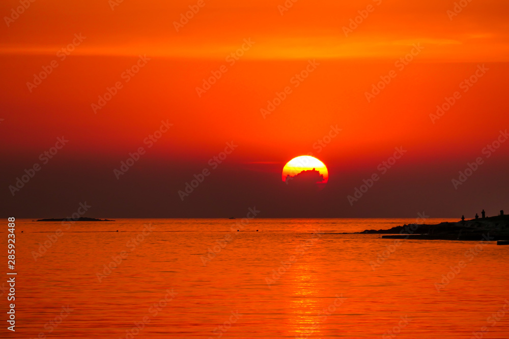 Romantic sunset by a beach. The sun sets over the horizon. The sun beams reflecting in the calm sea waters. There is an island on the side. Few birds flying around. The sky turns yellow and orange