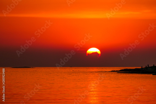 Romantic sunset by a beach. The sun sets over the horizon. The sun beams reflecting in the calm sea waters. There is an island on the side. Few birds flying around. The sky turns yellow and orange