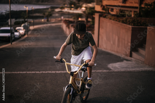 Trendy boy riding his bicycle on the warm light of a summer evening. Young man pedaling with BMX alone on the road near the sea. Teen that fatigue uphill on the bike. Youth recreation positive concept
