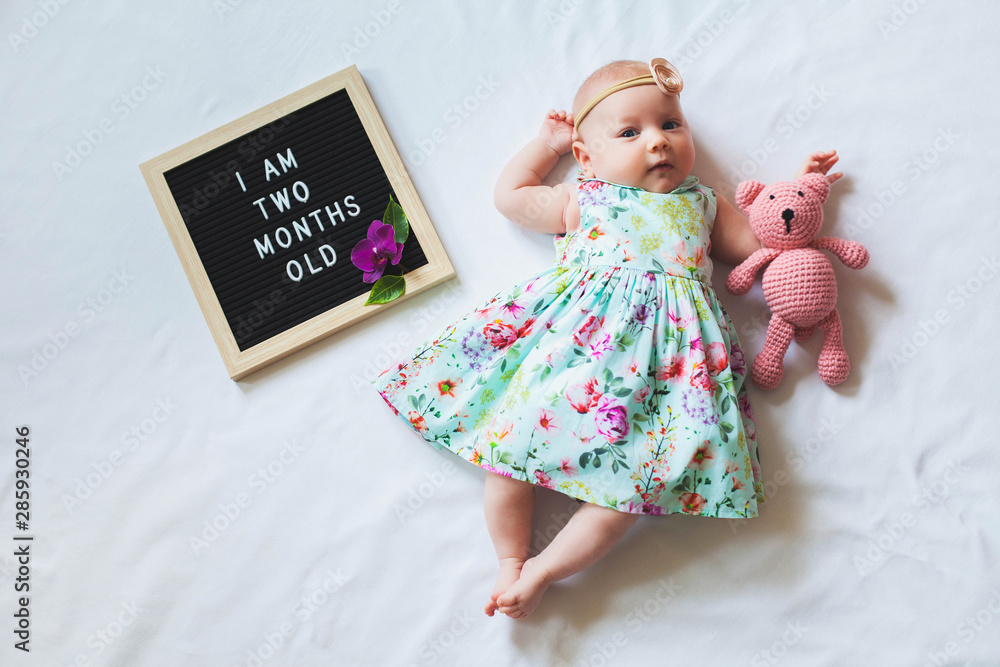 Φωτογραφία Two months old baby girl wearing floral dress laying down on white background and hugging knitted teddy bear