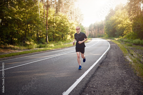Caucasian young curly man athlete runs sunny summer day on asphalt road in the forest. © Andrii