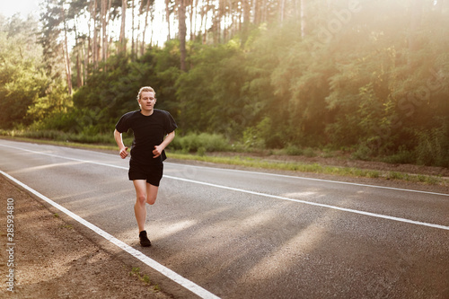 Caucasian young curly man athlete runs sunny summer day on asphalt road in the forest.