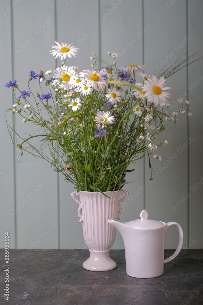 White teapot with tea and a bouquet of daisies and cornflowers in a