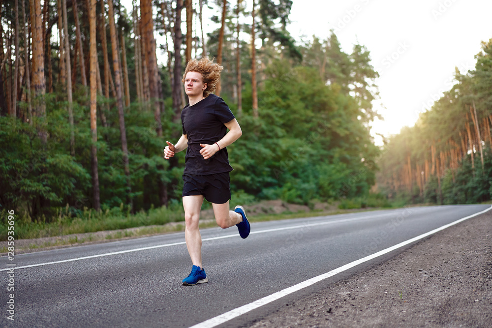 Caucasian young curly man athlete runs sunny summer day on asphalt road in the forest. Caucasian young curly man athlete runs sunny summer day on asphalt road in the forest.