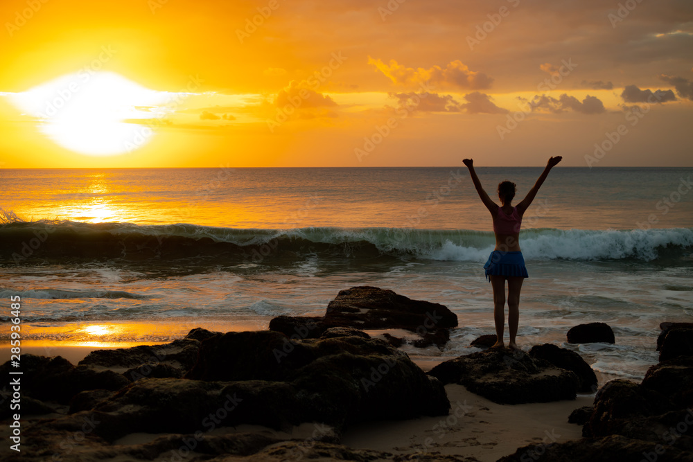 Excited young woman raising arms at the beach in front of the ocean. View from back. Sunset at the beach. Bali, Indonesia.