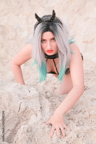 Young beautiful Caucasian woman in black swimwear with devil horns posing in desert landscape with sand.