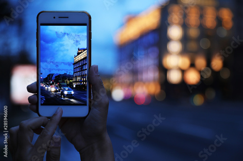 Woman taking photo of modern city in evening with smartphone, focus on hand. Bokeh effect