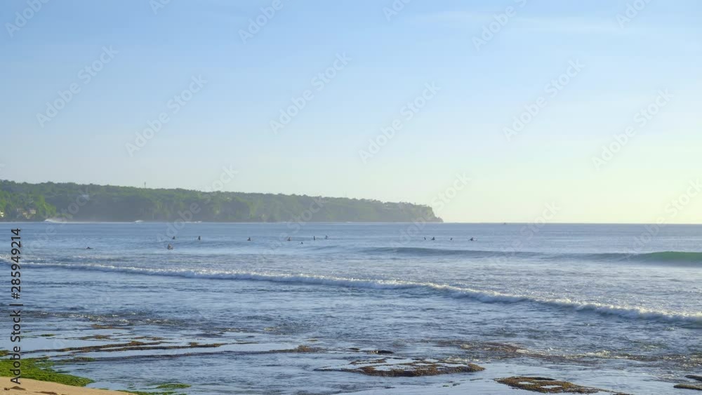 Surfers are swimming in blue water with waves during low tide near rocky beach in the evening Surfers are swimming in blue water with waves during low tide near rocky beach in the evening