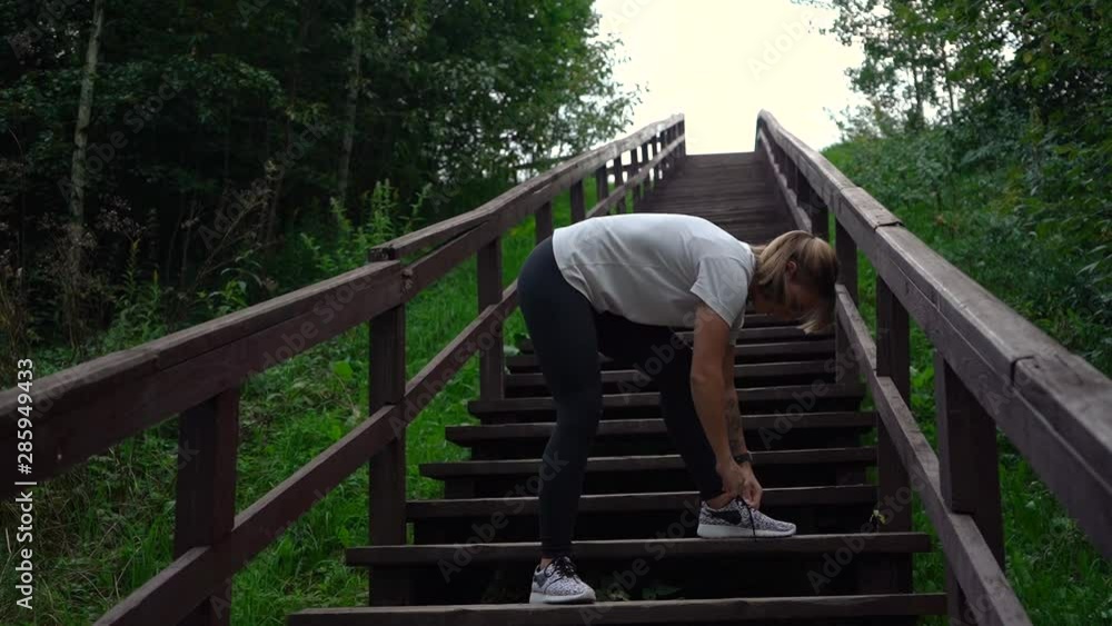 Pretty brunette senior fit female tying shoelace on sneakers on wood stairs in park. Slim woman in sportswear ties laces Pretty brunette senior fit female tying shoelace on sneakers on wood stairs in park. Slim woman in sportswear ties laces