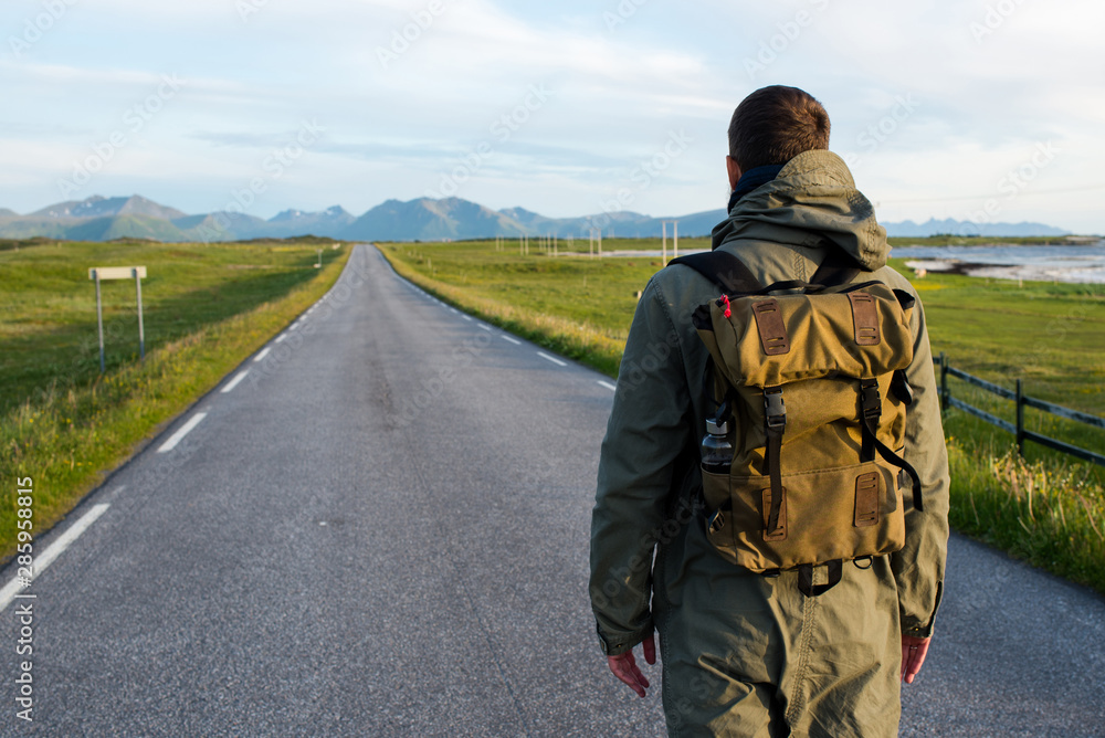 Fototapeta premium Man with backpack walking on an empty road. Amazing landscape, mountain and ocean. Scenic view. New way. Enjoy the moment, relaxation. Wanderlust. Travel, adventure, lifestyle. Explore North Norway
