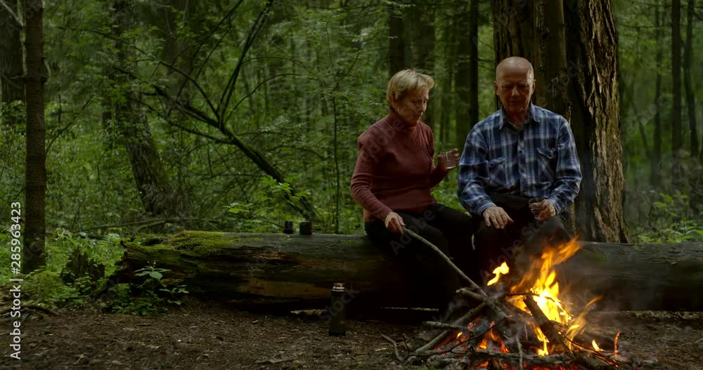 aged married couple are resting together in forest, looking on flaming fire aged married couple are resting together in forest, looking on flaming fire