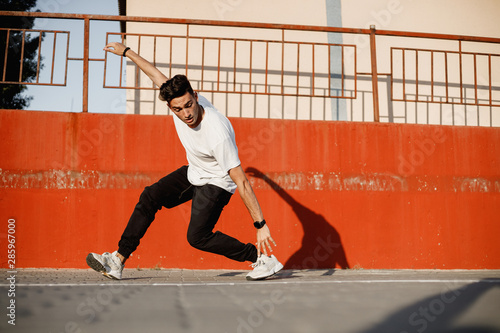 Stylish young guy dressed in jeans and white t-shirt is dancing brakedance in the street against a painted concrete wall in the sunny day