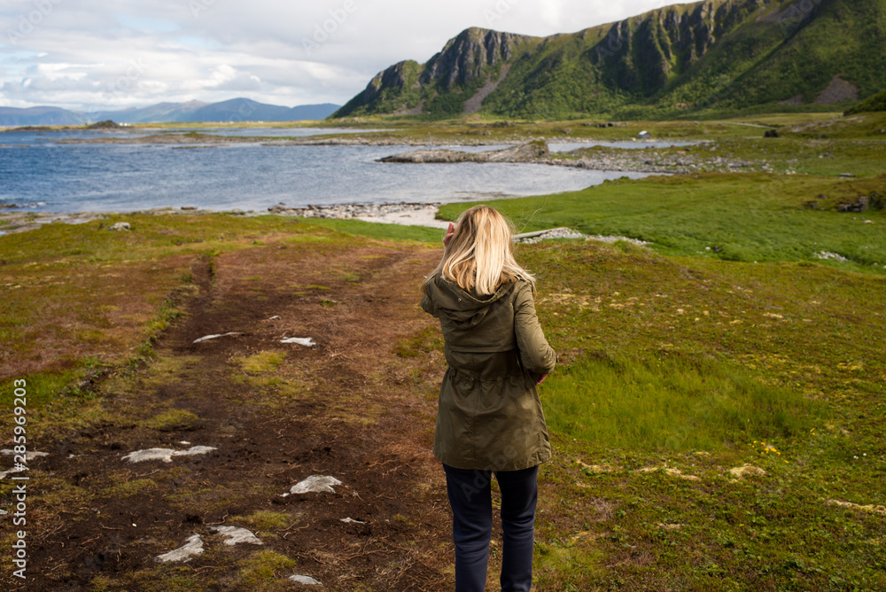 Naklejka premium Blond hair girl walking, looks forward and enjoys a stunning scenic view. Ocean and rocks, beautiful landscape. Wanderlust. Adventure, freedom, lifestyle. Explore North Norway. Summer in Scandinavia