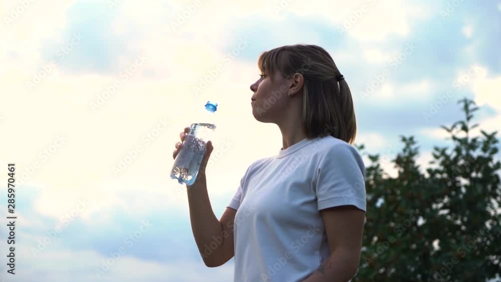 Young beautiful athletic girl in a white T-shirt is very tired and drinks water from a transparent bottle slow motion Young beautiful athletic girl in a white T-shirt is very tired and drinks water from a transparent bottle slow motion