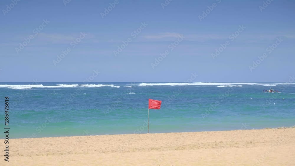 Slow motion of a red flag is swaying in wind on a tropical sandy beach during hot day with small boat in background Slow motion of a red flag is swaying in wind on a tropical sandy beach during hot day with small boat in background