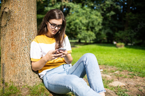 Young woman in blue jeans chatting via smartphone under park tree