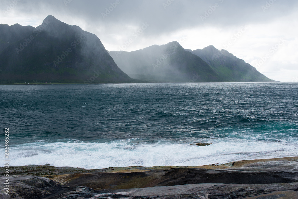 Beautiful nature landscape in North. Scenic outdoors view. Ocean with waves and mountains. Big dark stones. Dramatic storm clouds. Extreme weather, rain and wind. Explore Norway, summer adventure