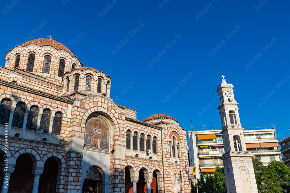 Cathedral Church of St. Nicholas, Volos. Stone Greek Orthodox Church on the town square with the city clock on the tower, among modern houses, summer, blue clear sky. Greece