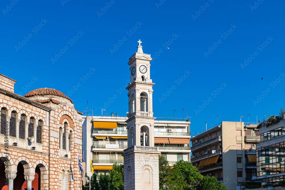 Cathedral Church of St. Nicholas, Volos. Stone Greek Orthodox Church on the town square with the city clock on the tower, among modern houses, summer, blue clear sky. Greece