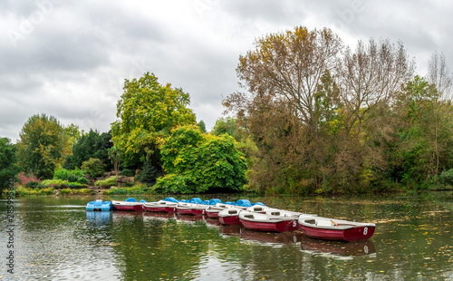 Panoramic view of a boating lake with paddle boats parked and park cafe outdoor tables, Battersea Park, London