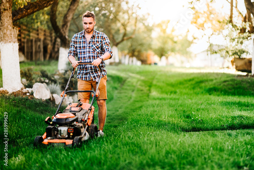 Εκτύπωση καμβά Portrait of young gardener using lawn mower for grass cutting in garden