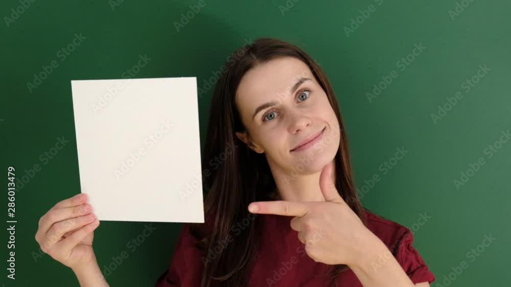 Portrait of beautiful smiling female wearing red t-shirt, posing with white blank paper with copy space for your advertisement information and looking at camera, against green background. Portrait of beautiful smiling female wearing red t-shirt, posing with white blank paper with copy space for your advertisement information and looking at camera, against green background.