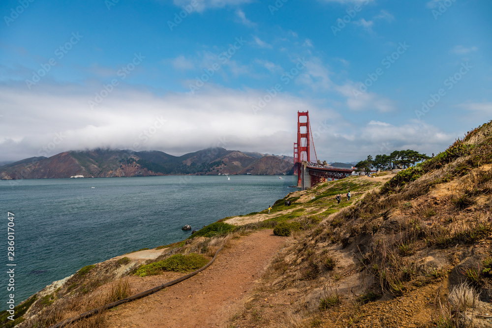 Golden Gate Bridge on the right with calm water from the San Francisco Bay on the left and a dirt footpath in the foreground Golden Gate Bridge on the right with calm water from the San Francisco Bay on the left and a dirt footpath in the foreground