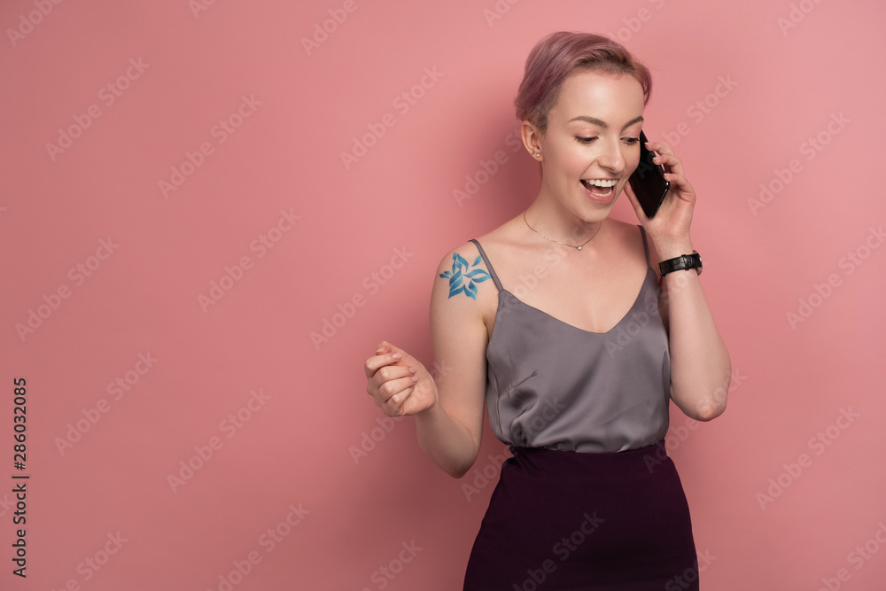 A girl with short pink hair and a tattoo in a gray top smiling speaks on the phone while standing on a pink background. A girl with short pink hair and a tattoo in a gray top smiling speaks on the phone while standing on a pink background.