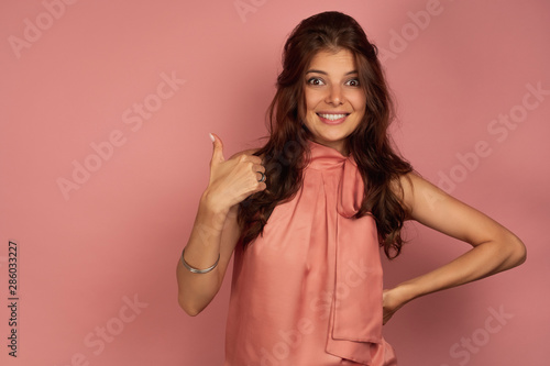 A girl in a pink top stands on a pink background, smiling embarrassed, showing a thumb up.