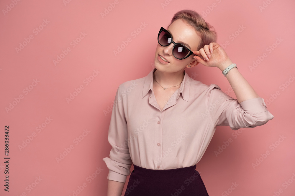 A girl with short pink hair in a blouse and skirt, smiling, looking at the camera in sunglasses, standing on a pink background. A girl with short pink hair in a blouse and skirt, smiling, looking at the camera in sunglasses, standing on a pink background.