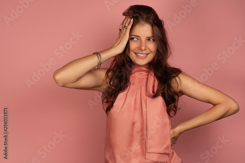 The brunette stands on a pink background in a dress and in embarrassment holds her head with her hand.