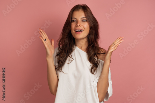 A dark-haired girl in a white top stands on a pink background laughing joyfully and raising her hands.