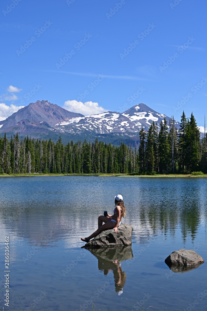 Young woman sitting on rock enjoys view of Middle and North Sisters volcanoes reflected in Scott Lake, Oregon on a calm sunny summer afternoon. Young woman sitting on rock enjoys view of Middle and North Sisters volcanoes reflected in Scott Lake, Oregon on a calm sunny summer afternoon.